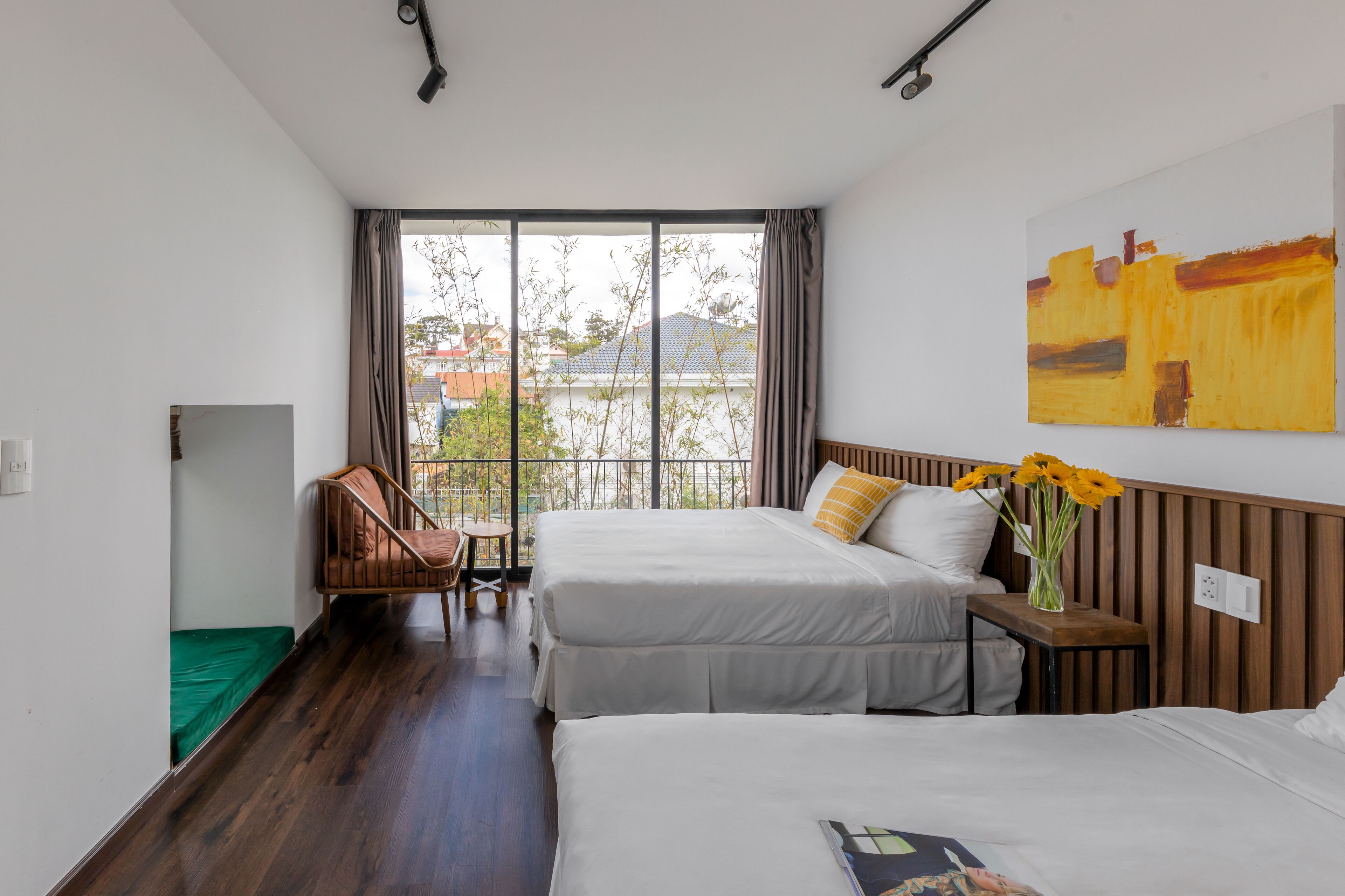A hotel room with two beds, a chair, a small table, and a large window offering a view of buildings and trees.