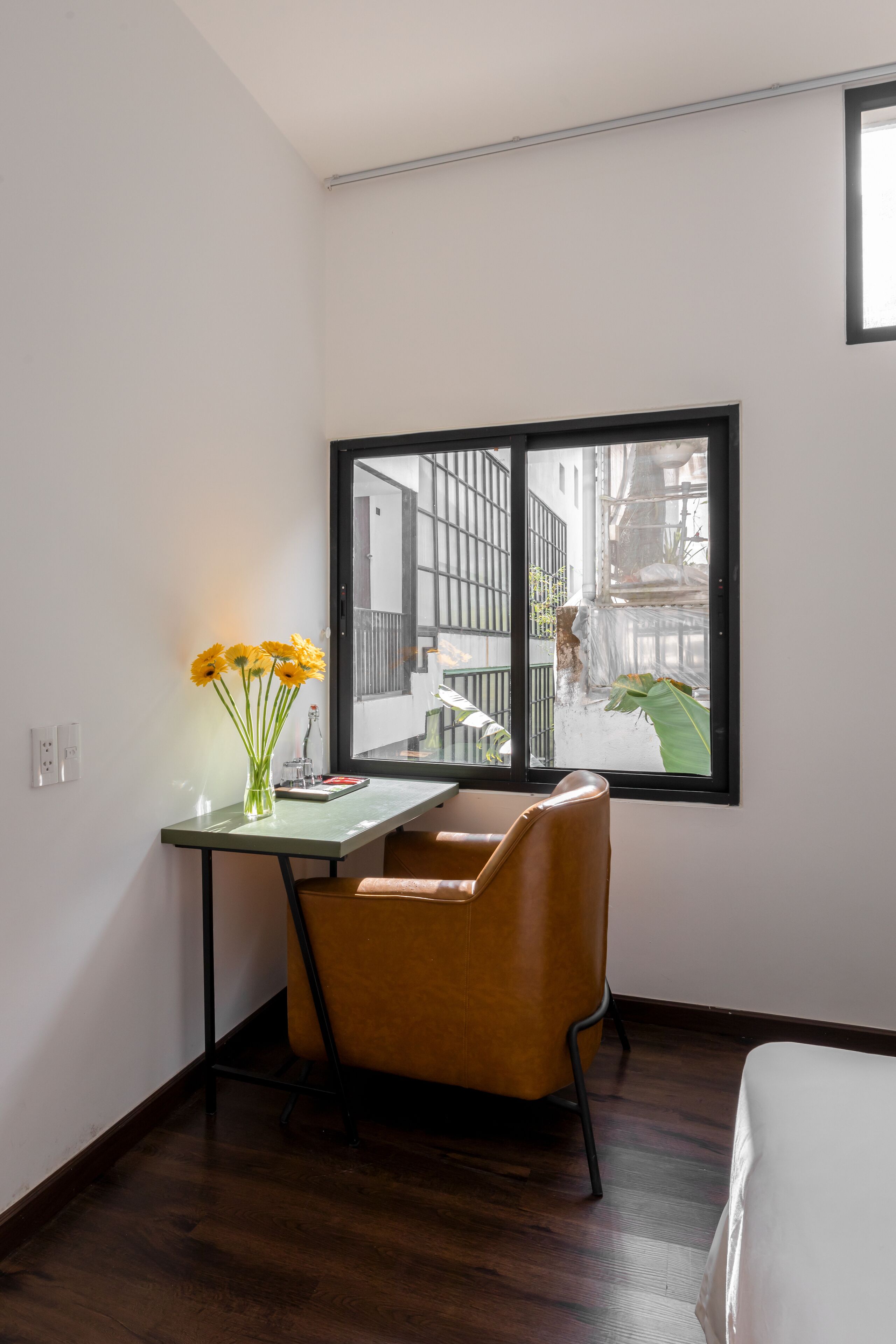 A corner room with a wooden floor, a desk with a vase of flowers, a chair, and a large window overlooking a street.