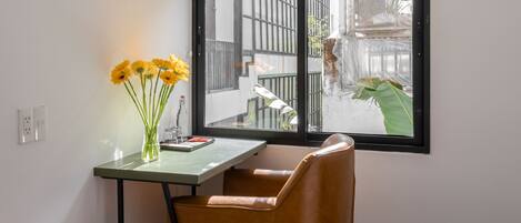A corner room with a wooden floor, a desk with a vase of flowers, a chair, and a large window overlooking a street.