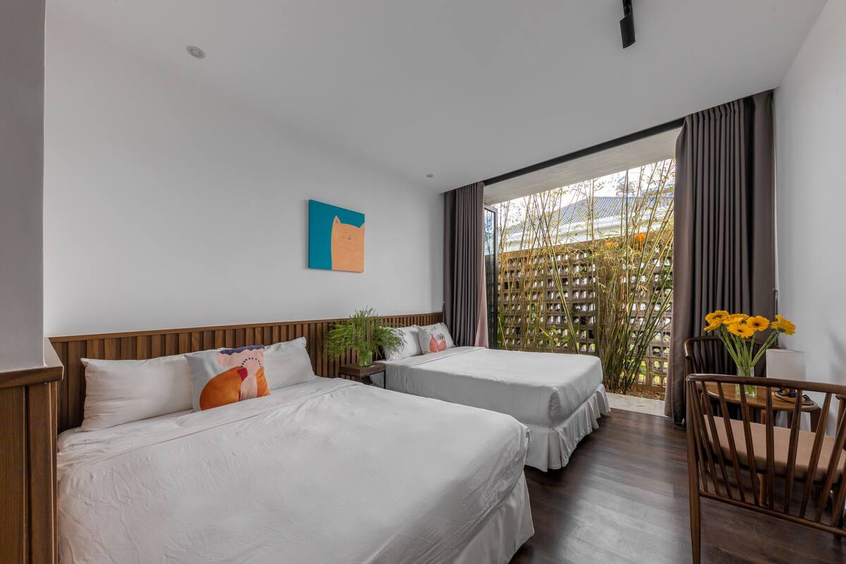 A hotel room with two beds, a wooden headboard, a small table with a chair, a potted plant, and a window with curtains.