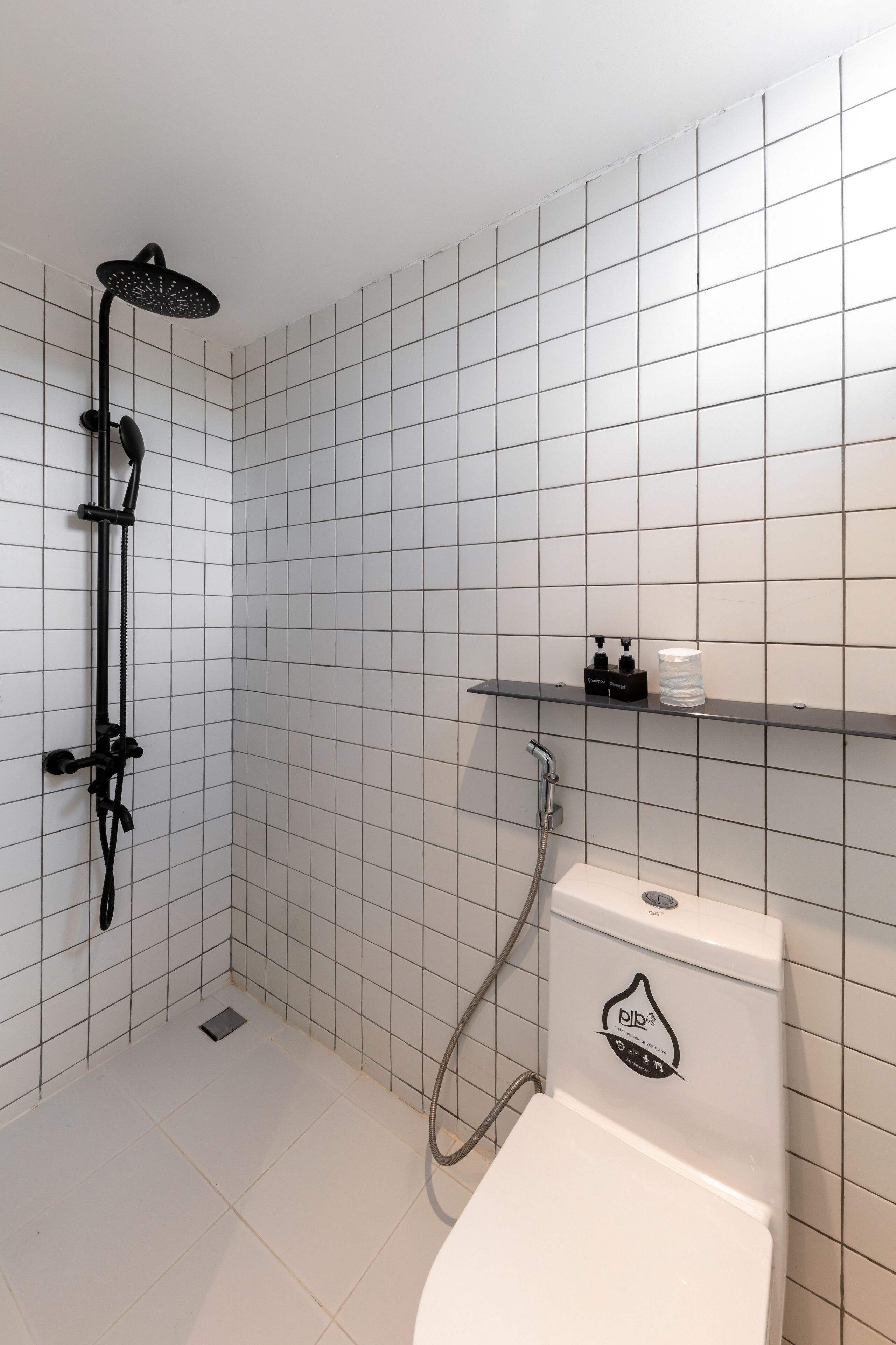 A modern bathroom with white tiled walls and floor, a black showerhead, a toilet with a water-saving label, and a shelf with toiletries.