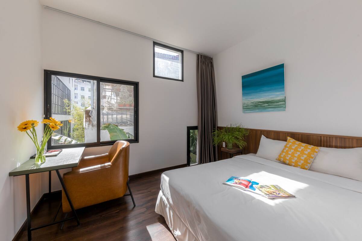A hotel room with a bed, a desk with a chair, a window with a view of buildings, and a painting on the wall.