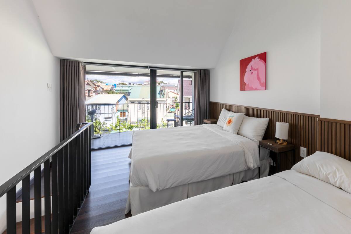 A hotel room with two beds, a balcony with a view of buildings, and a painting on the wall.