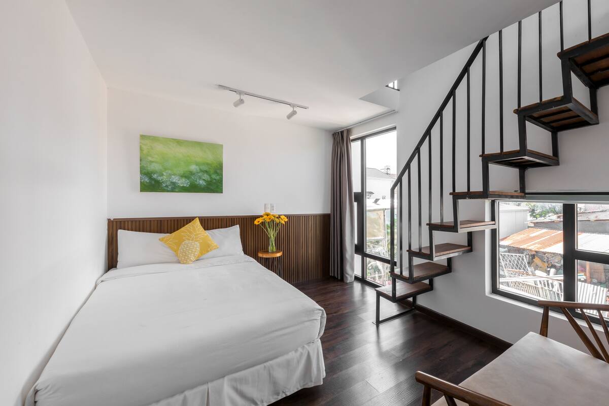 A modern hotel room with a large bed, a wooden headboard, a painting on the wall, a window with curtains, and a staircase with wooden steps.