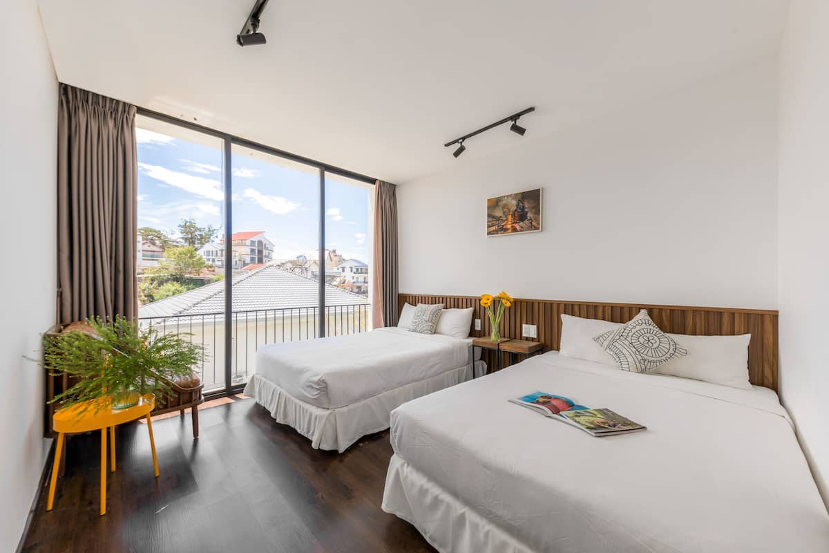 A hotel room with two beds, a large window with curtains, a small table with a plant, and a painting on the wall.