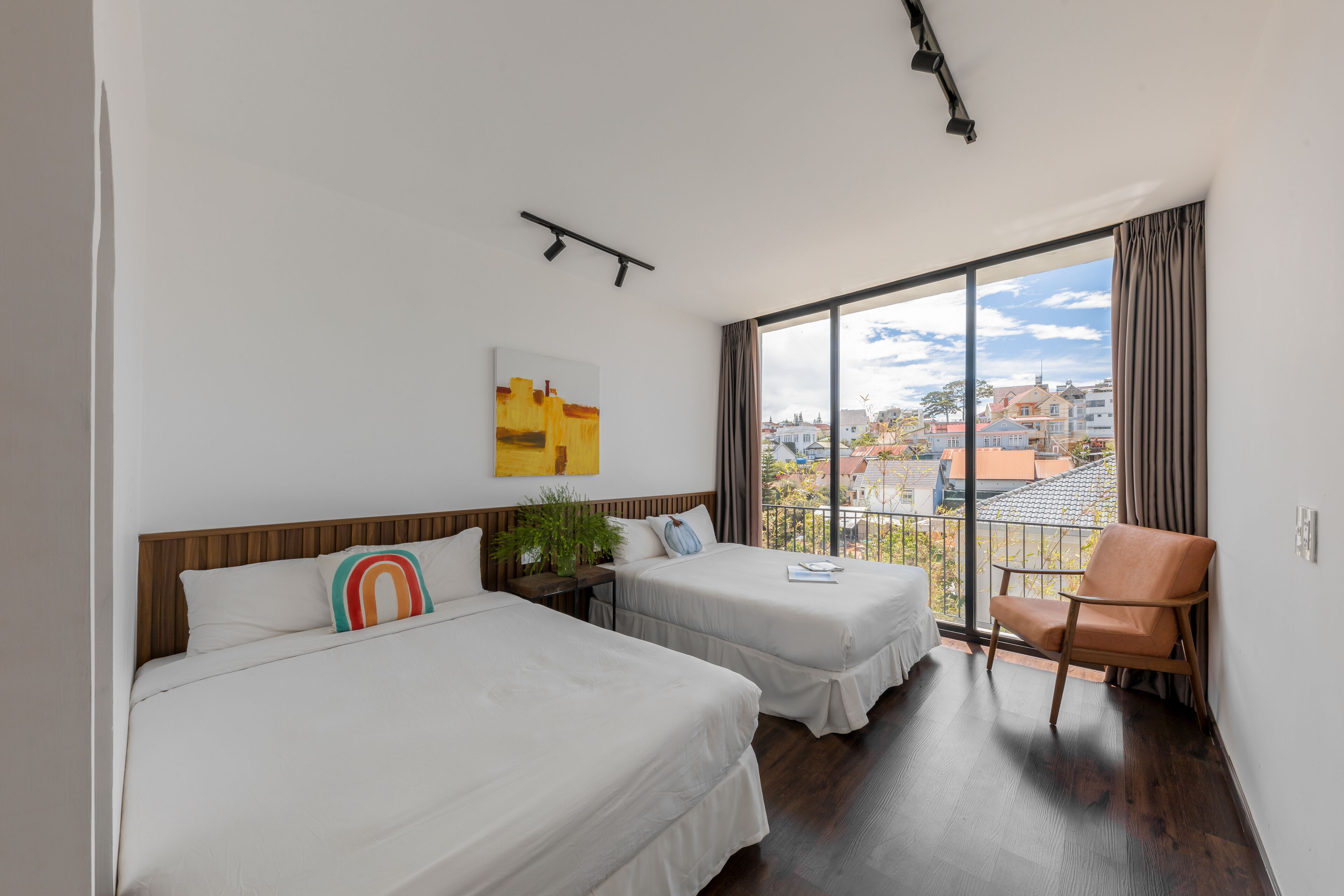 A hotel room with two beds, a large window with a view of buildings, a chair, and a plant.