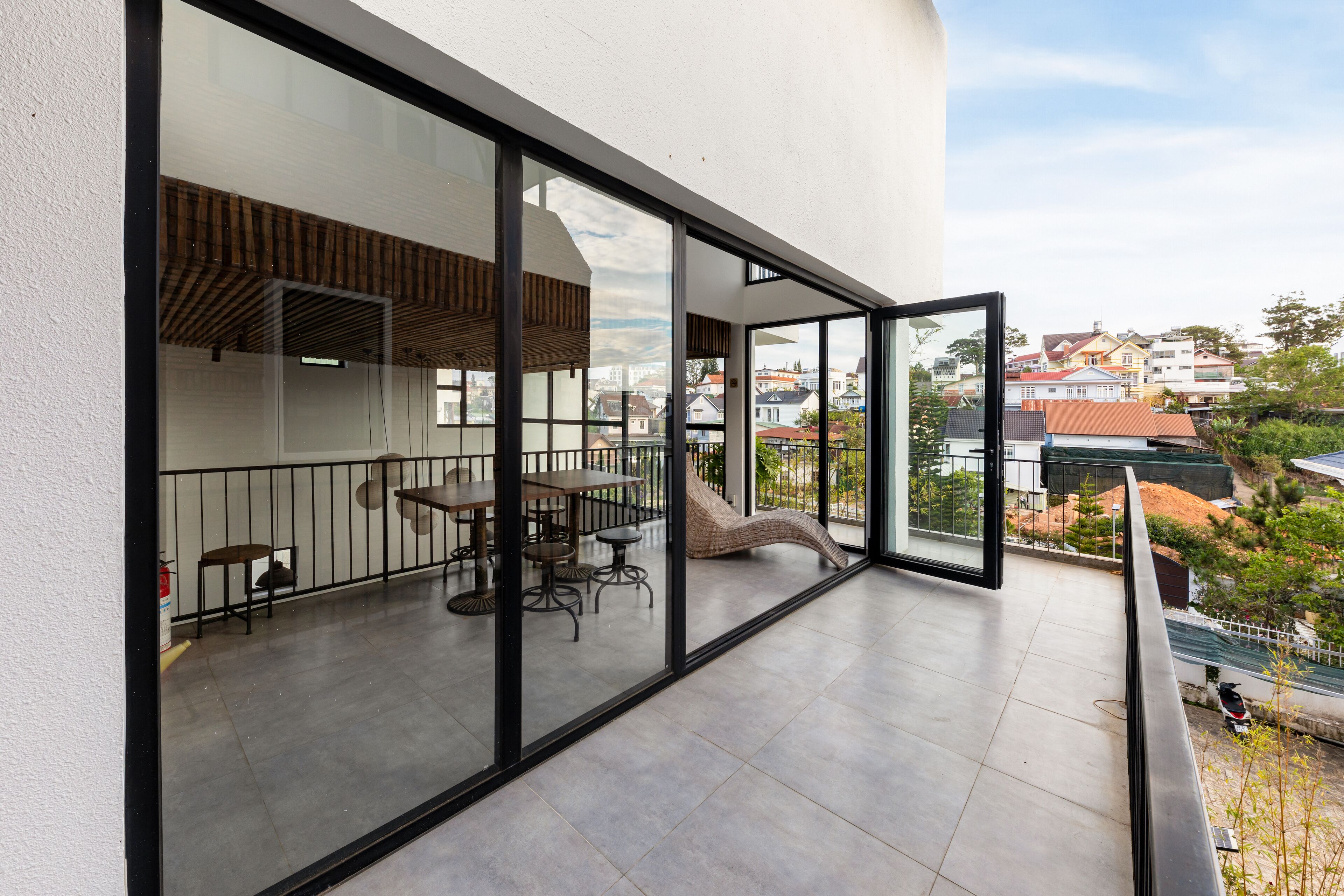 A modern balcony with glass doors, a dining area with tables and chairs, and a view of the cityscape.