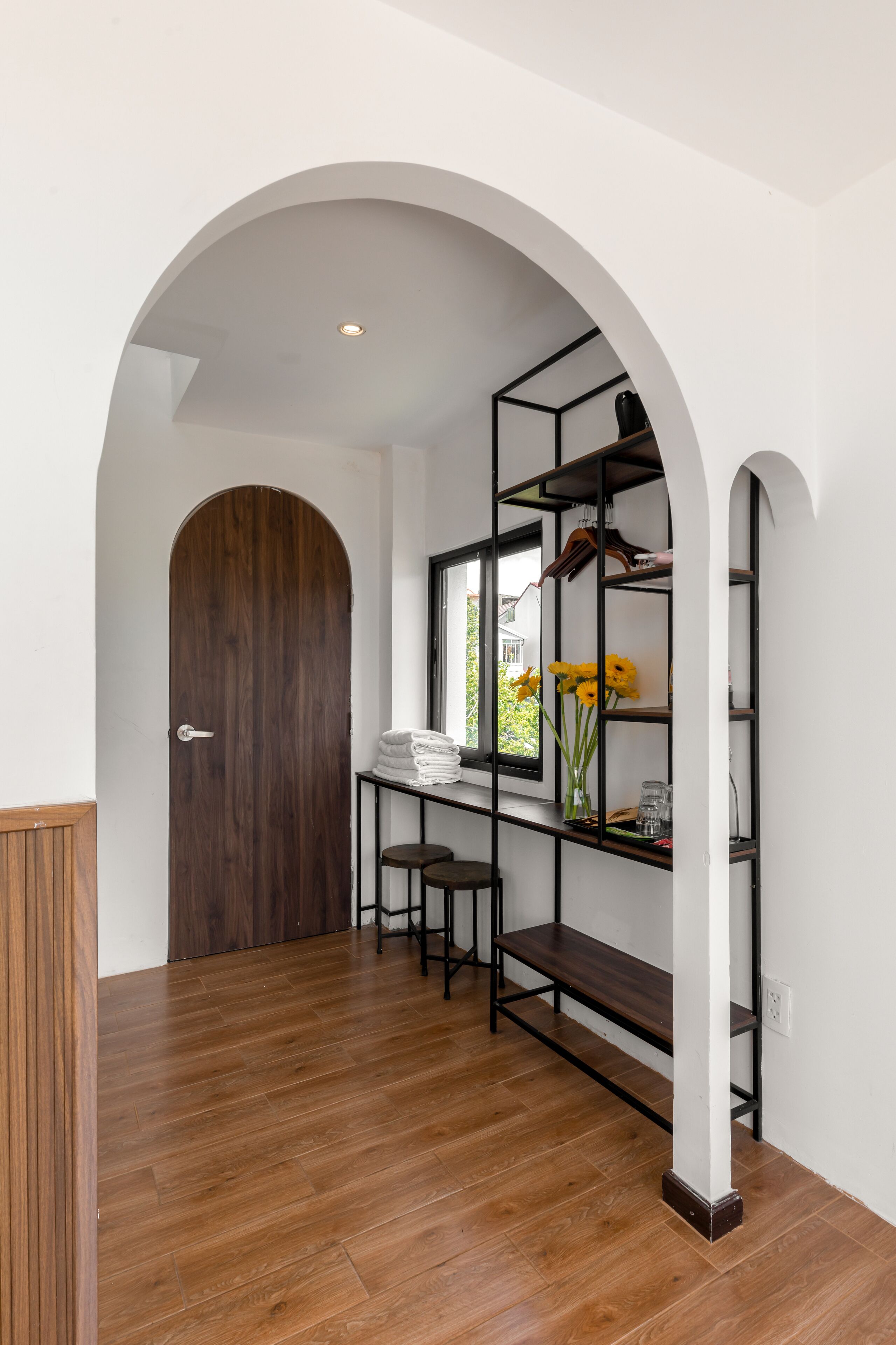 A hallway with a wooden door, a white wall, a black metal shelf unit with a window, and a bench.