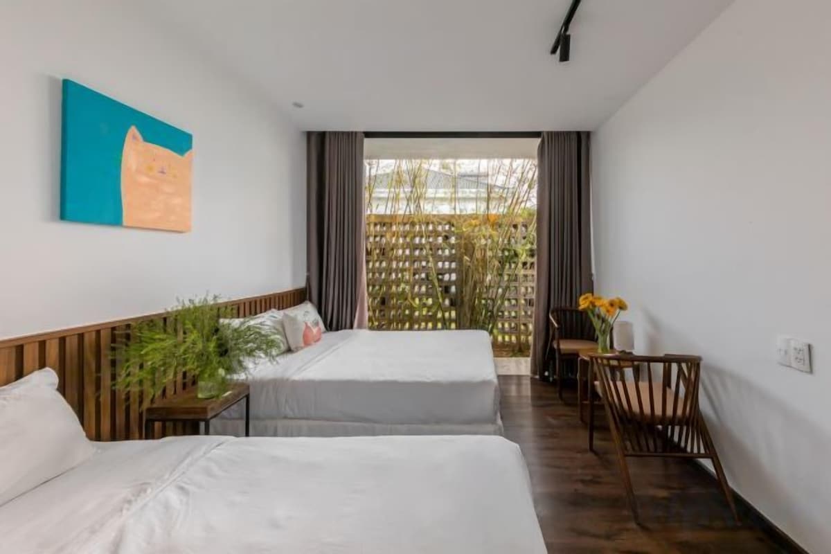 Habitación de hotel con dos camas, un cabecero de madera, una mesita con una planta y vista a una cerca de bambú a través de una ventana.