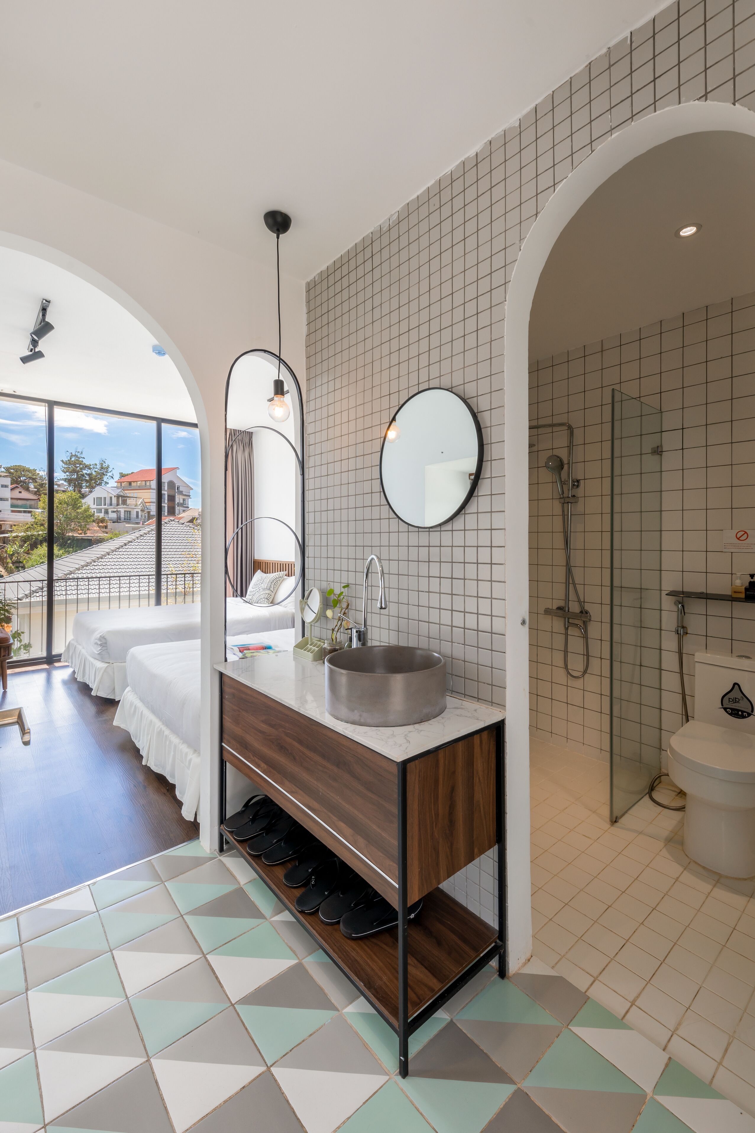 A modern bathroom with a large mirror, a sink, and a shower. There is a wooden vanity and a pair of beds visible through an arched doorway.