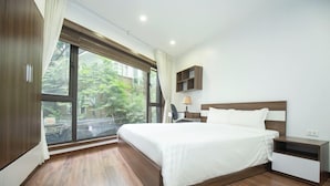 A modern bedroom with a large bed, a nightstand, a lamp, and a window with a view of greenery.