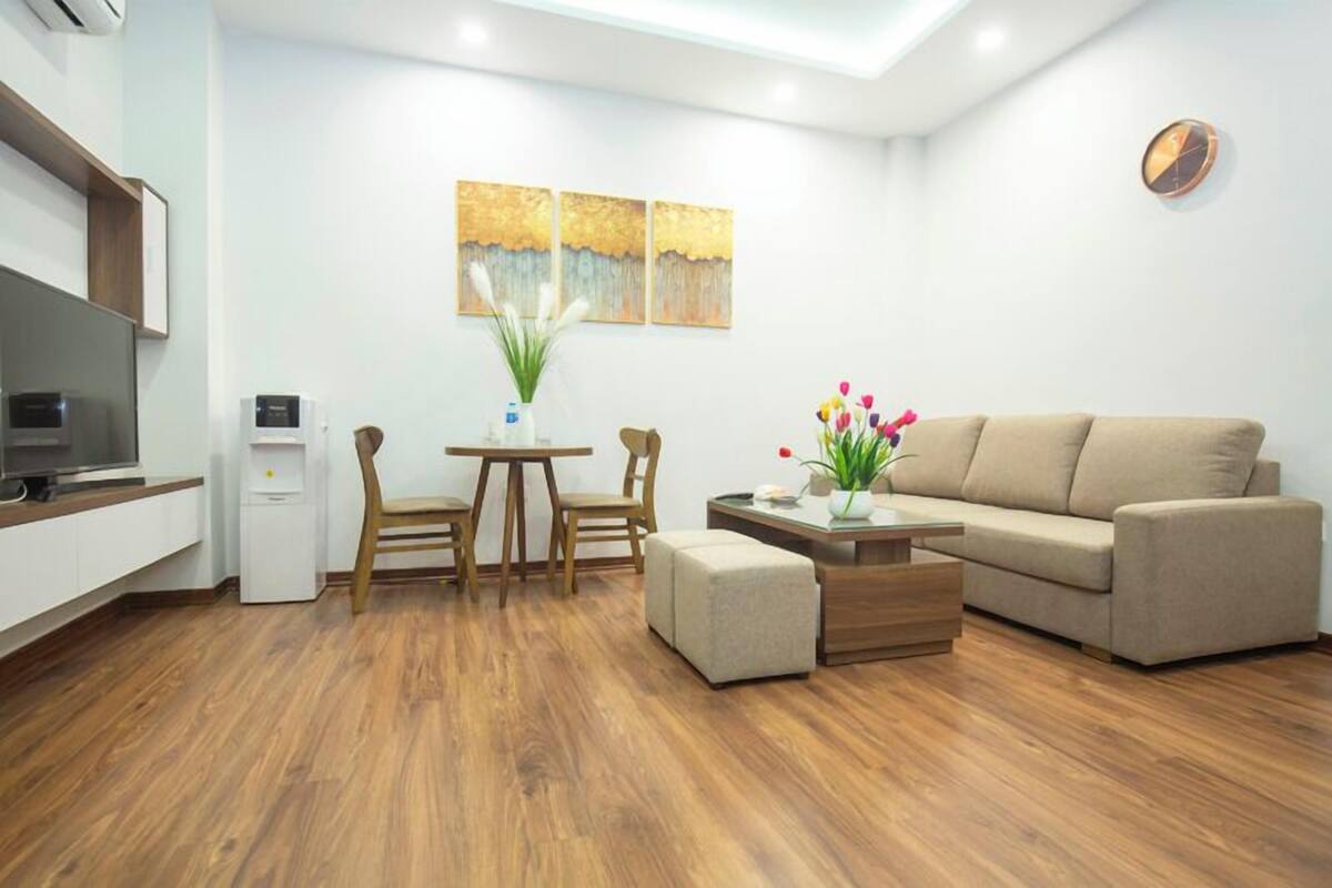 A modern living room with a sofa, ottoman, and a small table with a vase of flowers. There is a coffee machine and a TV on a stand.
