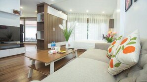 A modern living room with a sofa, a coffee table, and a television. There is a bookshelf and a window with curtains.