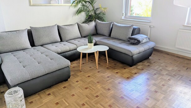 Un salon moderne avec un canapé d’angle, une petite table blanche, une plante en pot et une grande fenêtre.