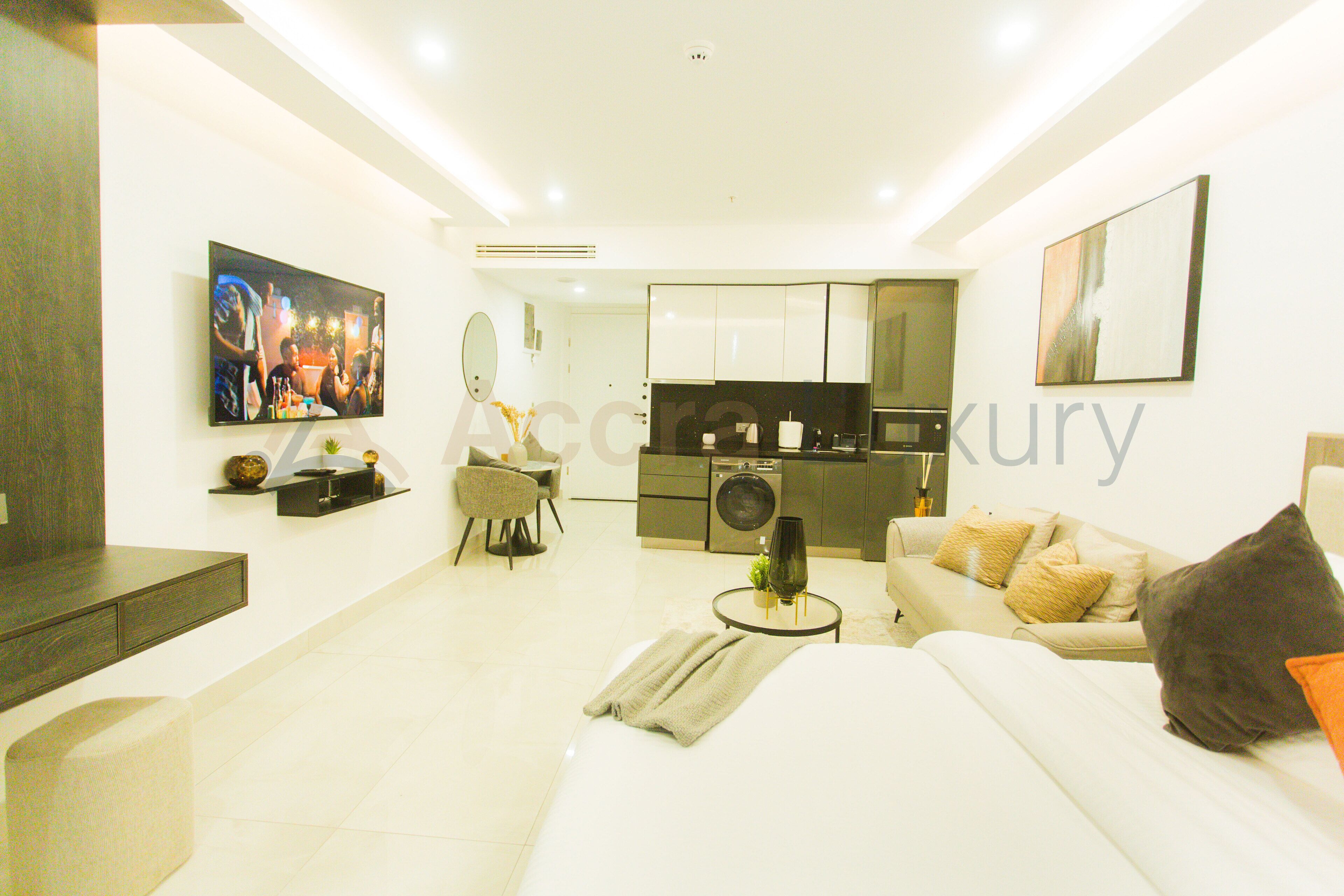 A modern living room with a sofa, a coffee table, and a TV. There is a kitchen area with cabinets and a washing machine.