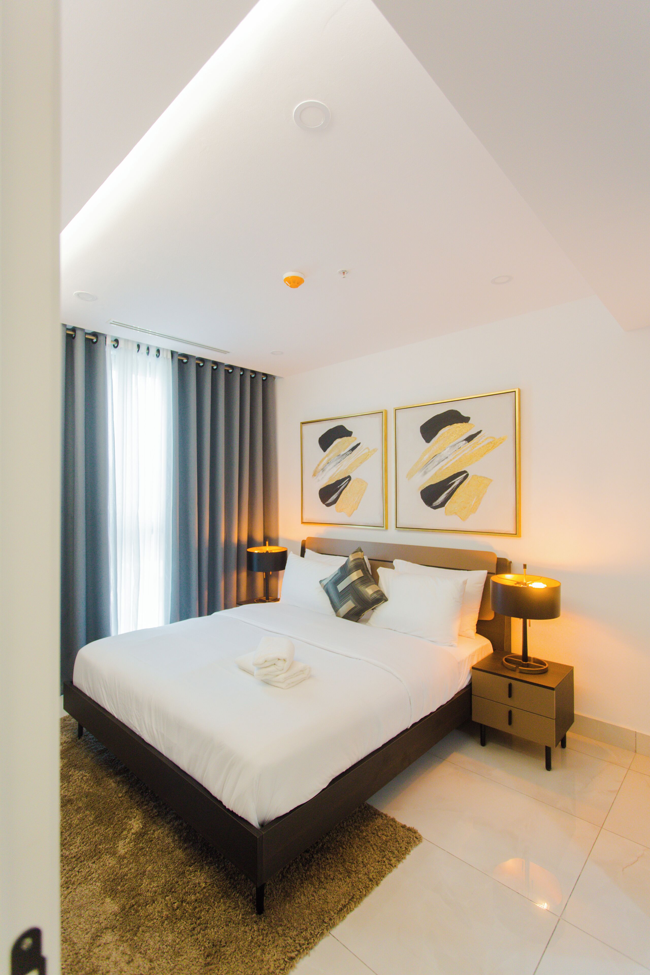 Une chambre d’hôtel moderne avec un grand lit, une table de chevet, deux lampes fixées au mur et une œuvre d’art abstraite accrochée au mur.