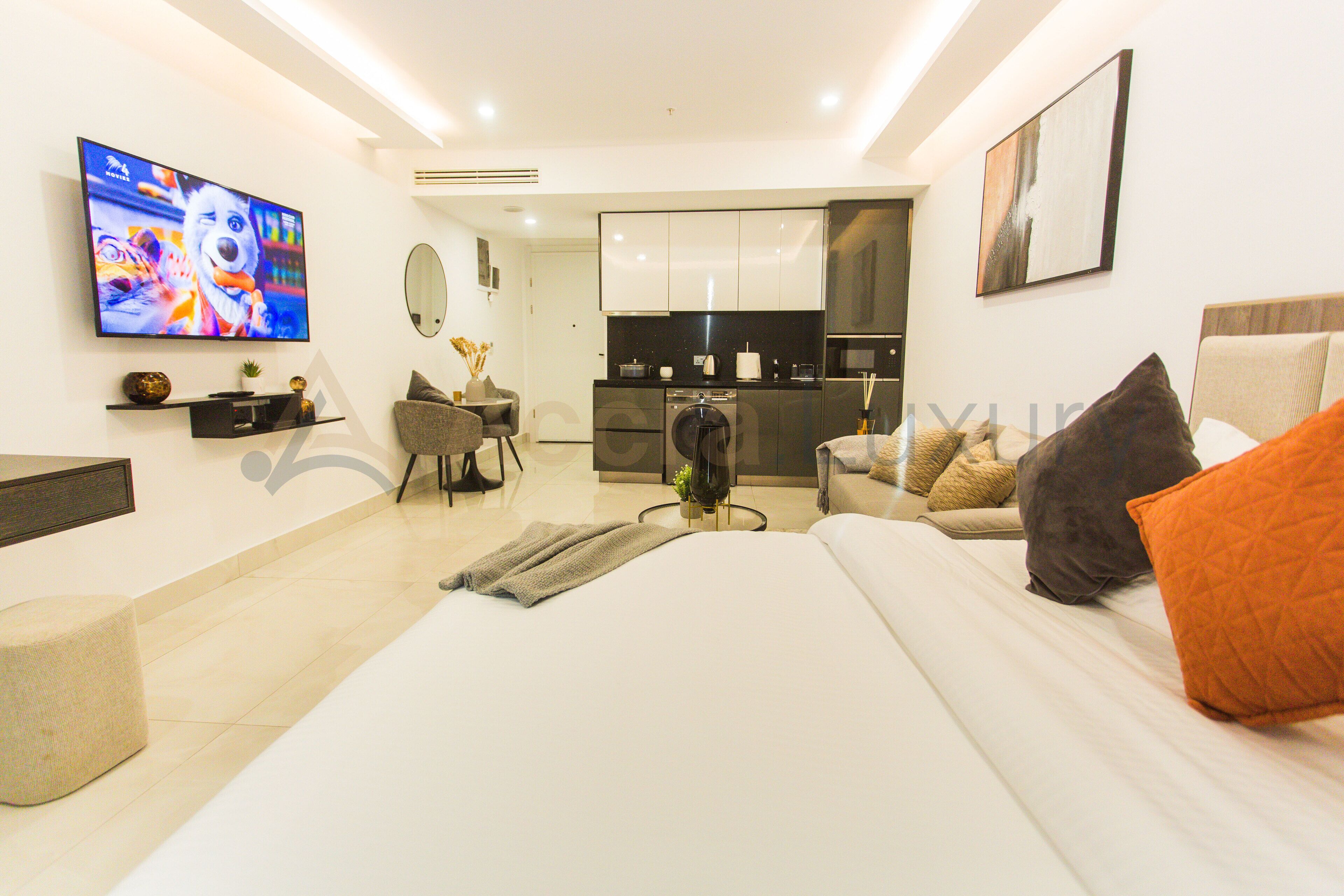 A modern hotel room with a large bed, a TV, a kitchen area, and a seating area.