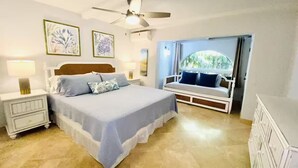 A bedroom with a bed, nightstand, dresser, ceiling fan, and a window with a view of greenery.