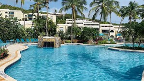 A swimming pool with a waterfall feature, surrounded by palm trees and lounge chairs.