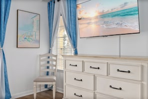 Una habitación con un tocador blanco, una silla, un ventanal con cortinas azules y un cuadro de una playa.