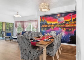 Un rincón comedor con una mesa dispuesta para cuatro personas, un mural con un paisaje urbano en la pared y vistas al exterior a través de amplios ventanales.