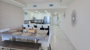 A spacious living area with a dining table, kitchen in the background, and a hallway leading to a door.