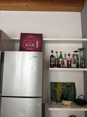 A kitchen with a refrigerator, shelves with liquor bottles, a decorative sign, and a teapot.