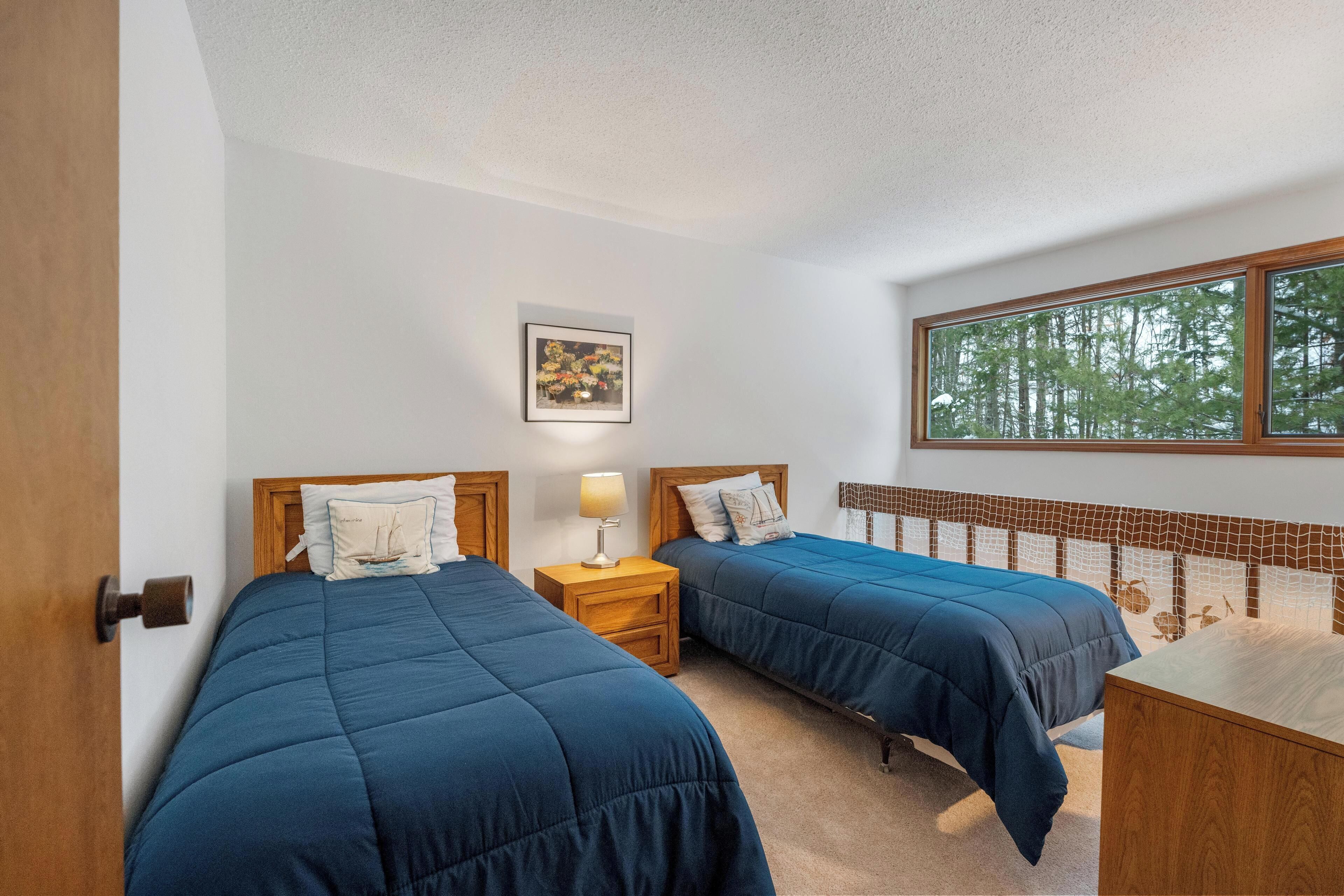 Une chambre avec deux lits, une table de chevet avec une lampe, une tête de lit en bois, une fenêtre donnant sur des arbres et un tableau au mur.