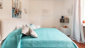 Een slaapkamer met een bed, kussens, een nachtkastje, een kleine tafel en een raam met gordijnen.