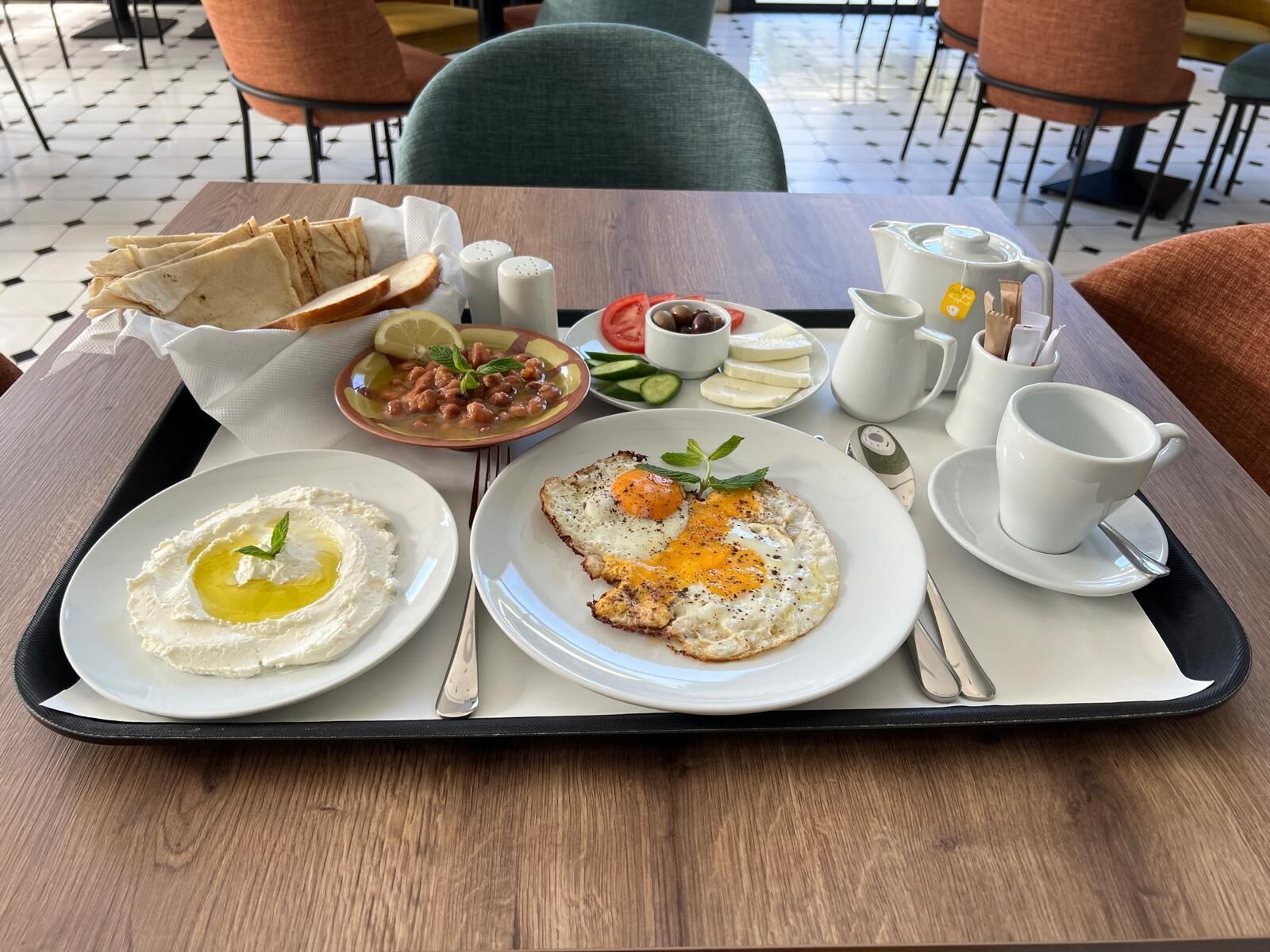 A tray with a plate of eggs, a plate of hummus, a plate of beans, a jug of water, a cup of coffee, and a small bowl of olives.