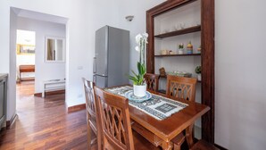 A dining area with a wooden table, chairs, and a decorative plant. A refrigerator and a shelf with kitchenware are also visible.