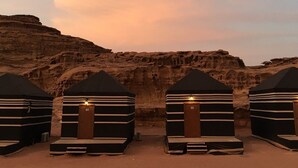 Cuatro tiendas a rayas negras y blancas, con puerta de madera y una lámpara, situadas en un paisaje desértico con un risco rocoso al fondo.