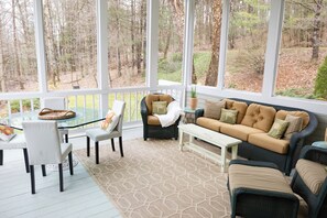 Relax in this serene outdoor space surrounded by a wooded view, featuring a glass-top table, comfortable seating, and a cozy sofa.
