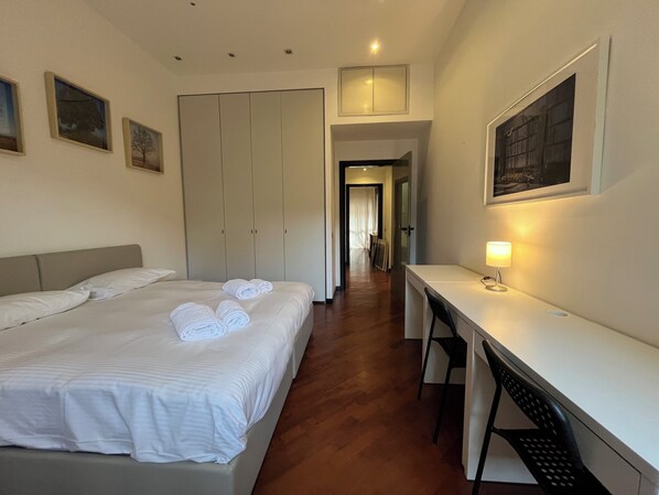 Quarto de hotel com cama, escrivaninha e cadeira. Quadros emoldurados na parede e uma luminária na escrivaninha.