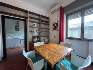 Cozinha compacta com armários brancos, pia e bancada pequena. Há uma mesa de jantar com quatro cadeiras, uma TV em um suporte e uma janela com cortina vermelha.