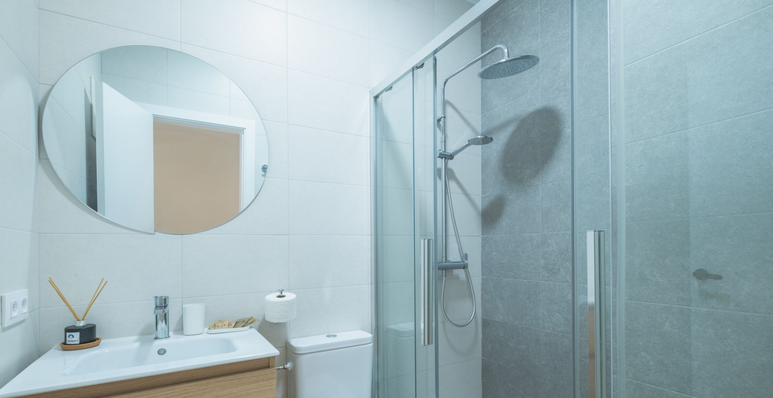 Une salle de bain moderne avec une douche à parois de verre, un miroir rond et un lavabo blanc avec un meuble en bois.