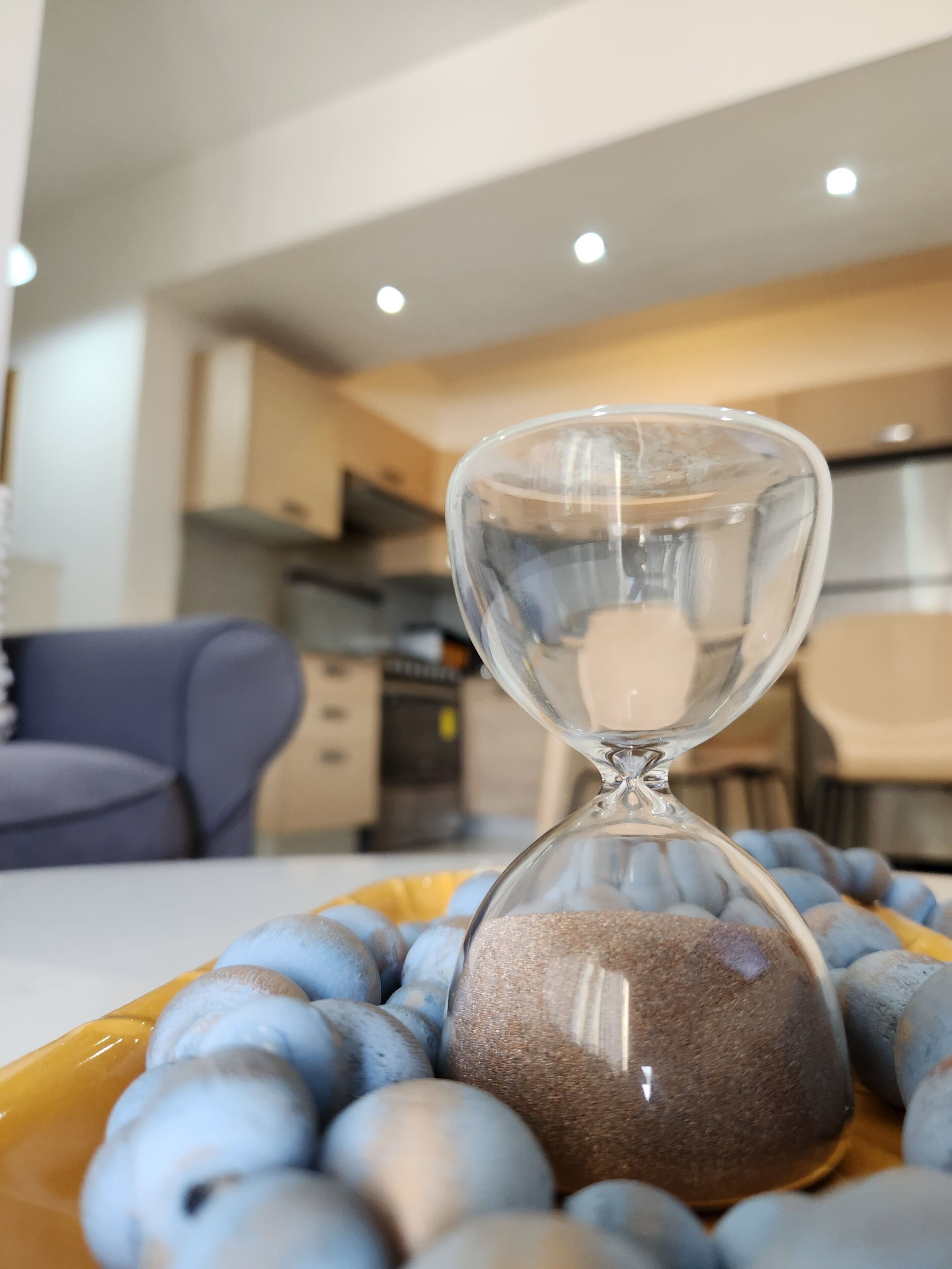 An hourglass surrounded by blue objects on a table in a modern kitchen with cabinets and a dining area in the background.