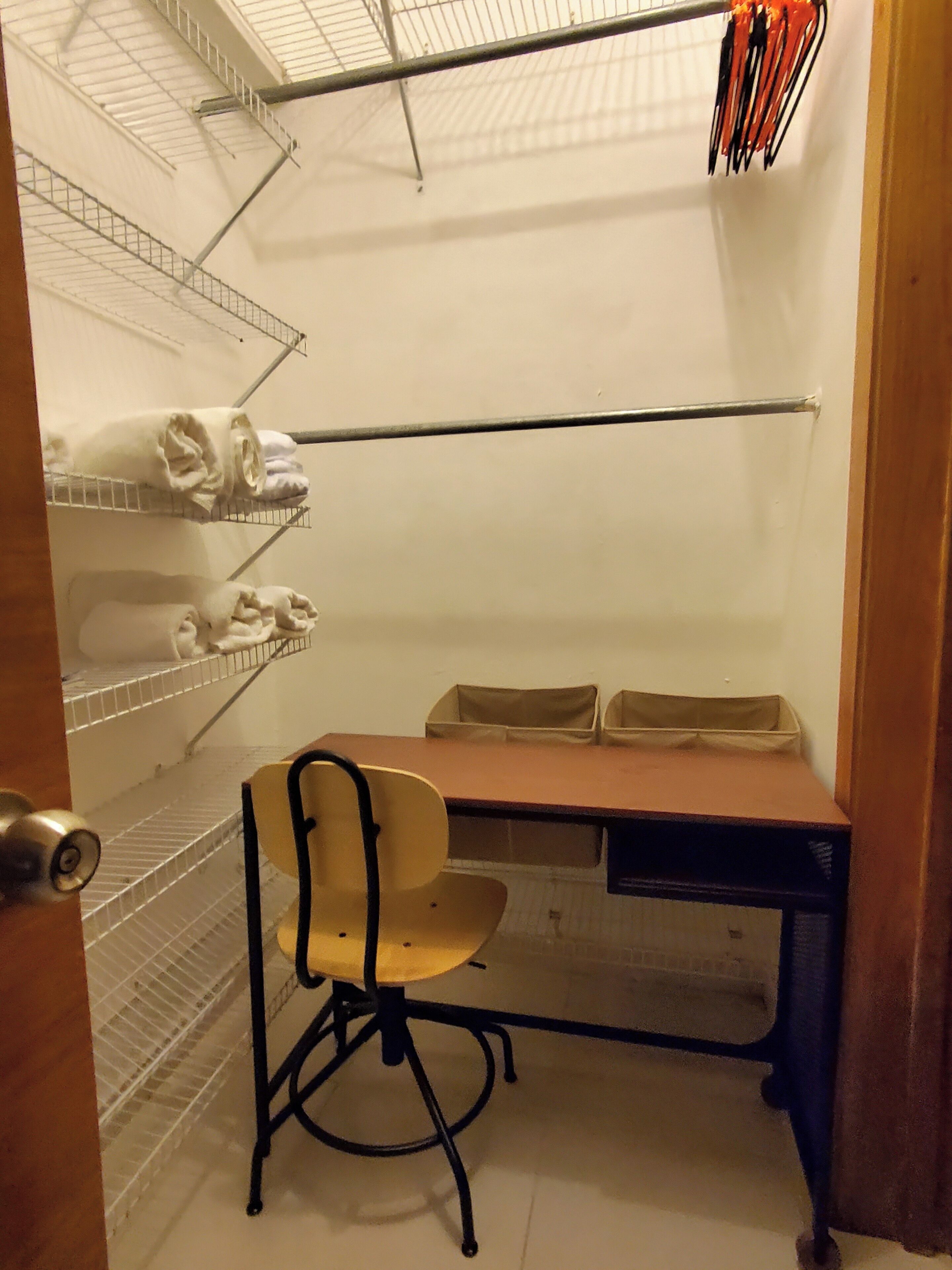 Ein kleiner Büroraum mit Schreibtisch, Stuhl, Regalen mit gefalteten Handtüchern und einem Metallgestell mit orangefarbenen und schwarzen Gegenständen.