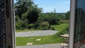 Een kamer met een schuifdeur die uitkomt op een tuin met een grindpad, begroeiing en een houten tafel met stoelen.