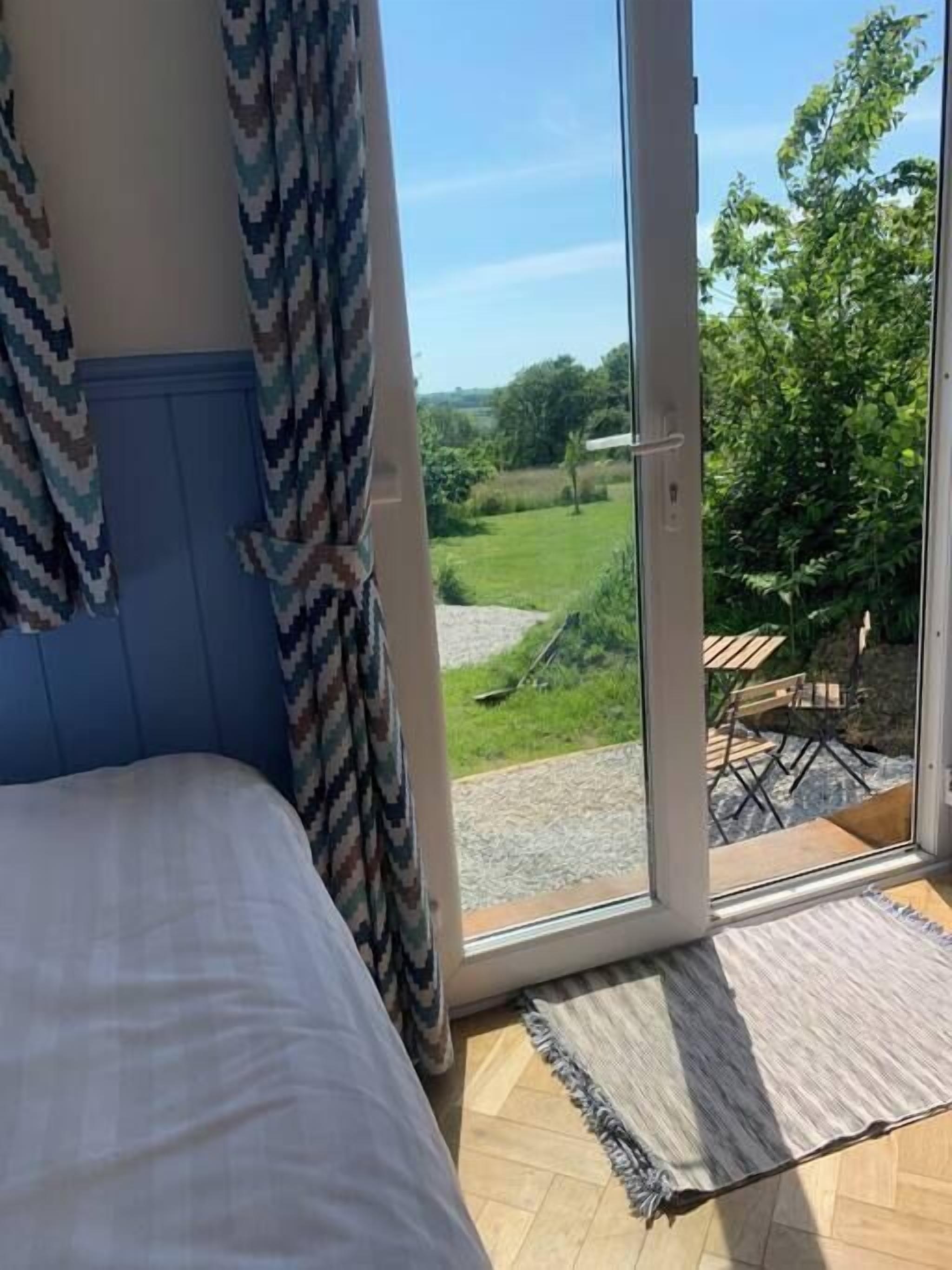 A bedroom with a bed, a view of a garden through a sliding glass door, and a small outdoor seating area with chairs.