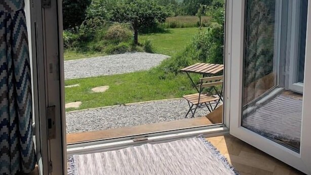 Een kamer met een grote glazen schuifdeur die uitkomt op een tuin met een grindpad, een houten tafel en een stoel.