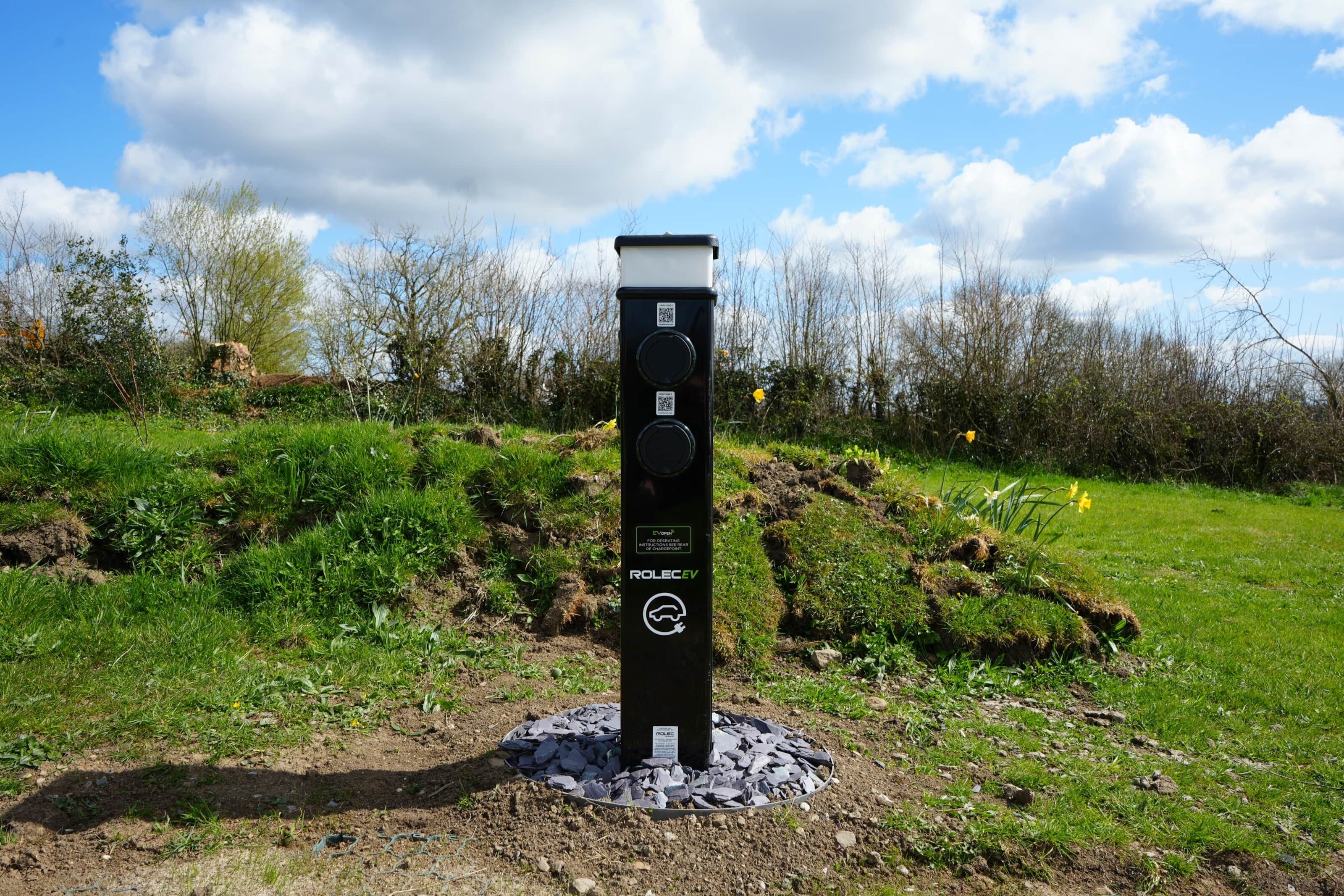 A black electric vehicle charging station with QR codes and the text "ROLEC EV" in a grassy outdoor area with trees and a clear sky.