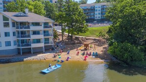 Une vue sur un immeuble à plusieurs étages avec des balcons, une plage de sable avec des chaises longues et une aire de jeux, et un petit bateau sur l’eau.