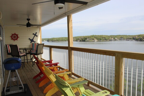 Eine überdachte Terrasse mit bunten Adirondack-Stühlen, einem Deckenventilator und Blick auf einen See mit Gebäuden in der Ferne.