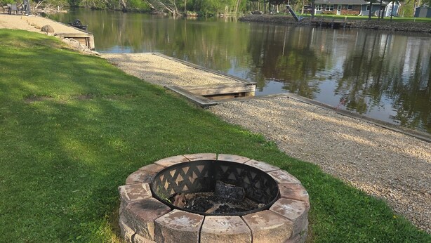 A circular stone fire pit surrounded by grass, a wooden dock extending into a calm lake, and trees in the background.