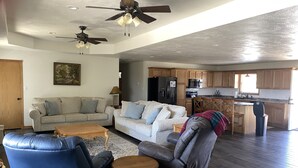 A spacious living room with a sofa, chairs, and a coffee table. There are ceiling fans, a kitchen with wooden cabinets, and a refrigerator.