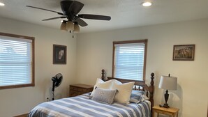 A bedroom with a bed, a ceiling fan, a nightstand with a lamp, and two windows with blinds.