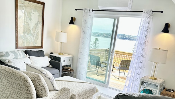 Una sala de estar con un sofá estampado, un sillón negro, una mesita con una lámpara, y vistas a un balcón con piso de madera y sillas.