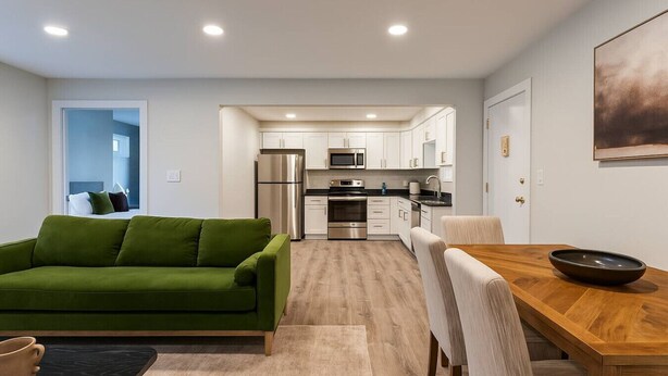 A modern apartment with a green sofa, a wooden dining table, and a kitchen in the background.