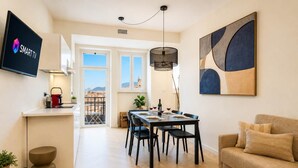 Un espace de vie moderne comprenant un coin repas, une cuisine avec un évier apparent, un téléviseur à écran plat et une grande fenêtre offrant une vue sur la ville.